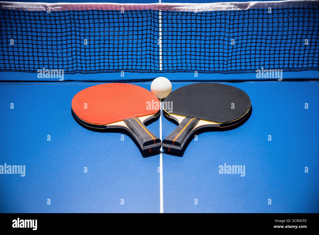 Schwarz und rot Tischtennisschläger und eine weiße Kugel auf der blauen Tischtennisplatte mit einem Netz, zwei Tischtennispaddel ist ein Sportwettkampfgerät i Stockfoto