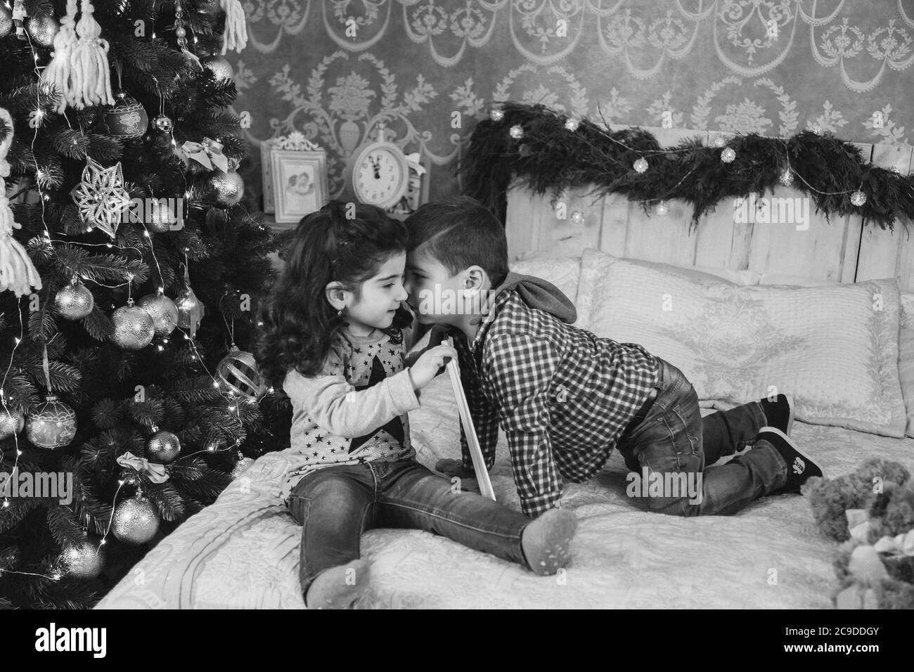 Weihnachtsporträt von kleinen Kindern, die auf dem Bett mit Geschenken unter dem weihnachtsbaum sitzen. Junge küsst das Mädchen. Winterurlaub Weihnachten und Neujahr Stockfoto