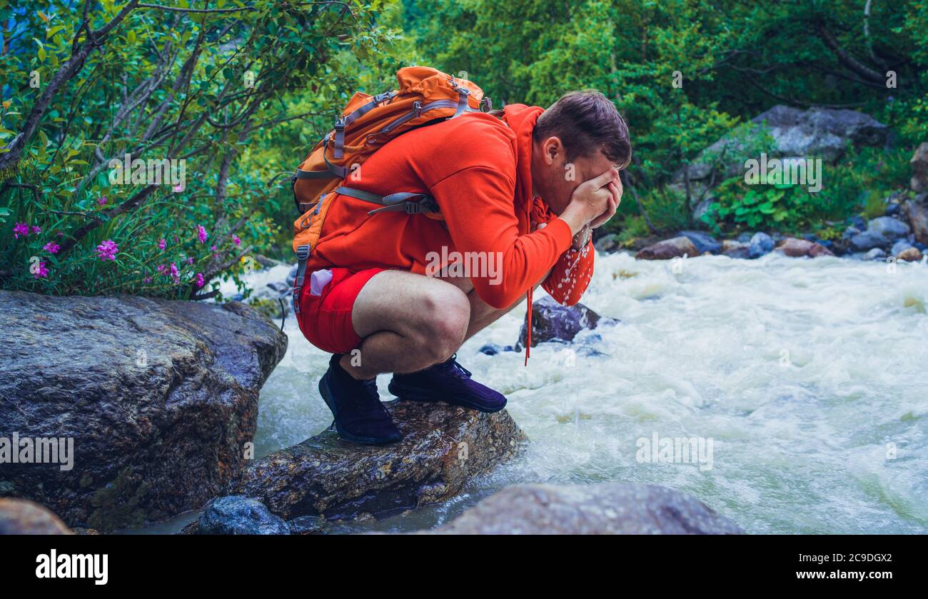 Männlicher Reisender wäscht sein Gesicht mit Bergwasser. Mann Tourist sitzt auf Felsen in der Nähe des Bergflusses und erfrischt sich. Stockfoto