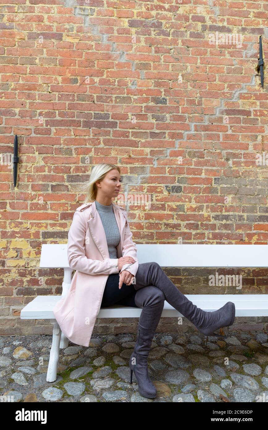 Junge schöne blonde Frau sitzt auf Holzbank gegen Backsteinmauer Stockfoto