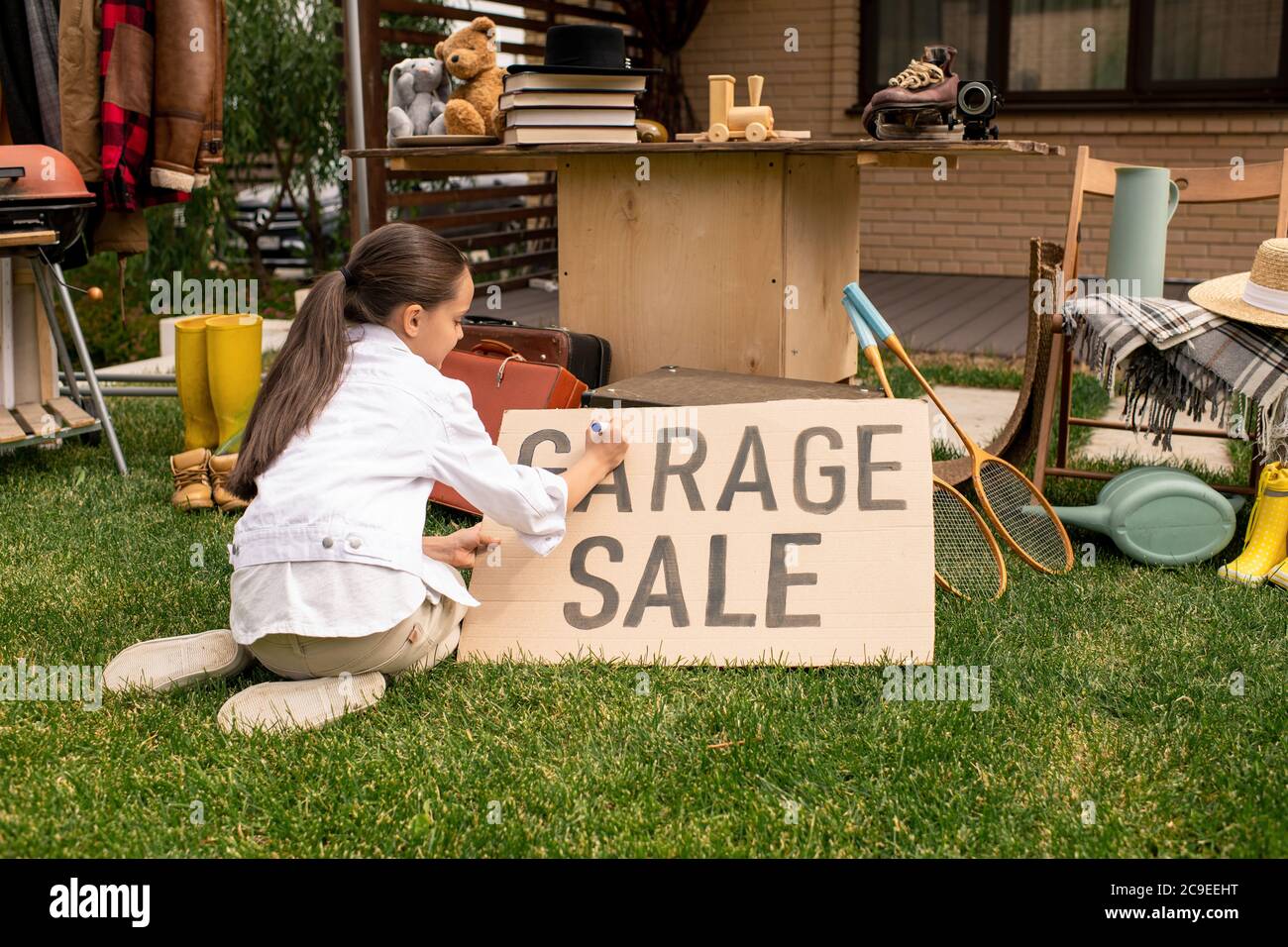 Rückansicht der Brünette Mädchen auf Gras sitzen und schreiben Garage Verkauf auf Banner im Hinterhof Stockfoto
