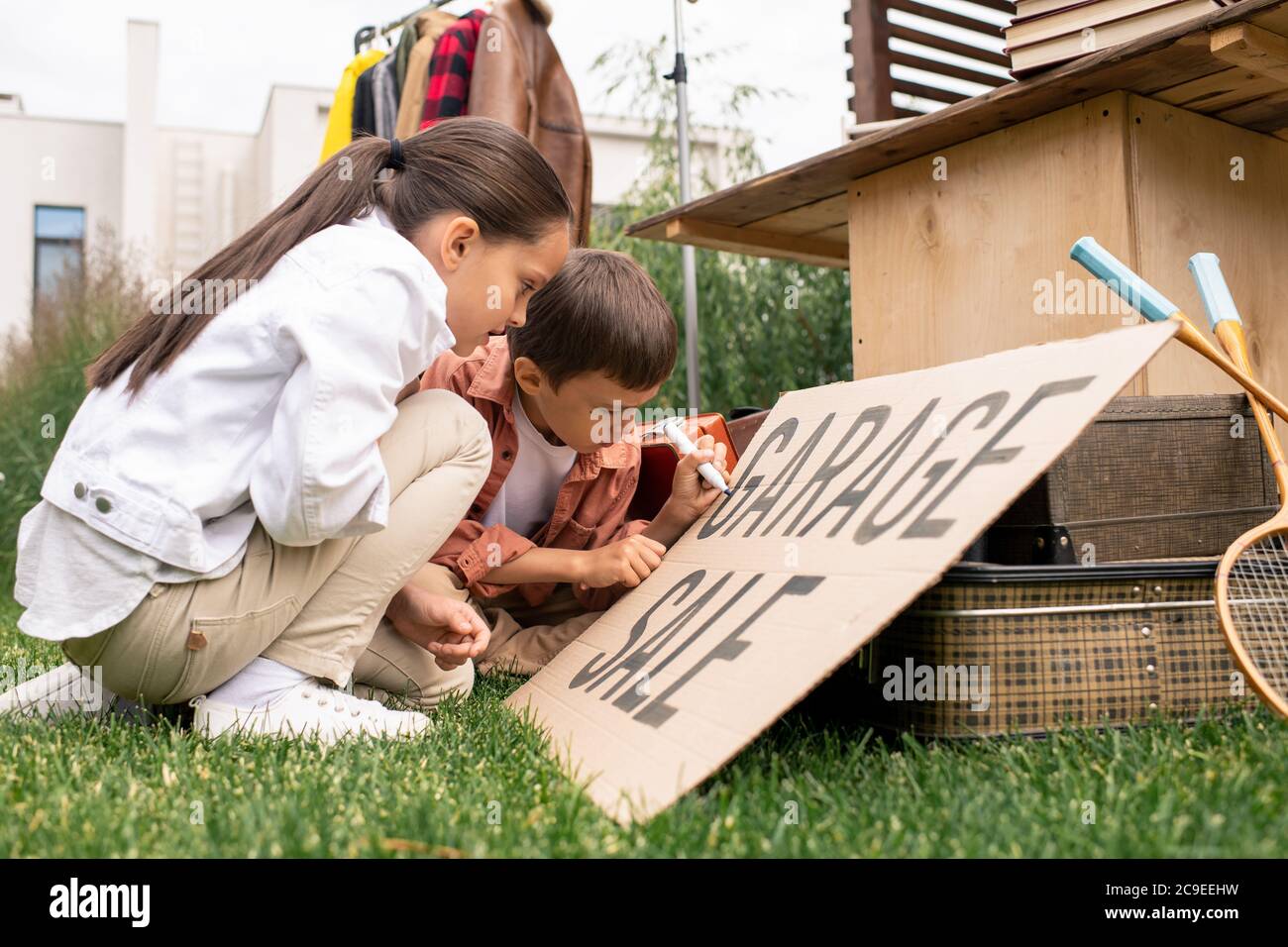 Kreative niedliche Kinder sitzen auf Gras und machen Garage Verkauf Zeichen zusammen im Hinterhof Stockfoto