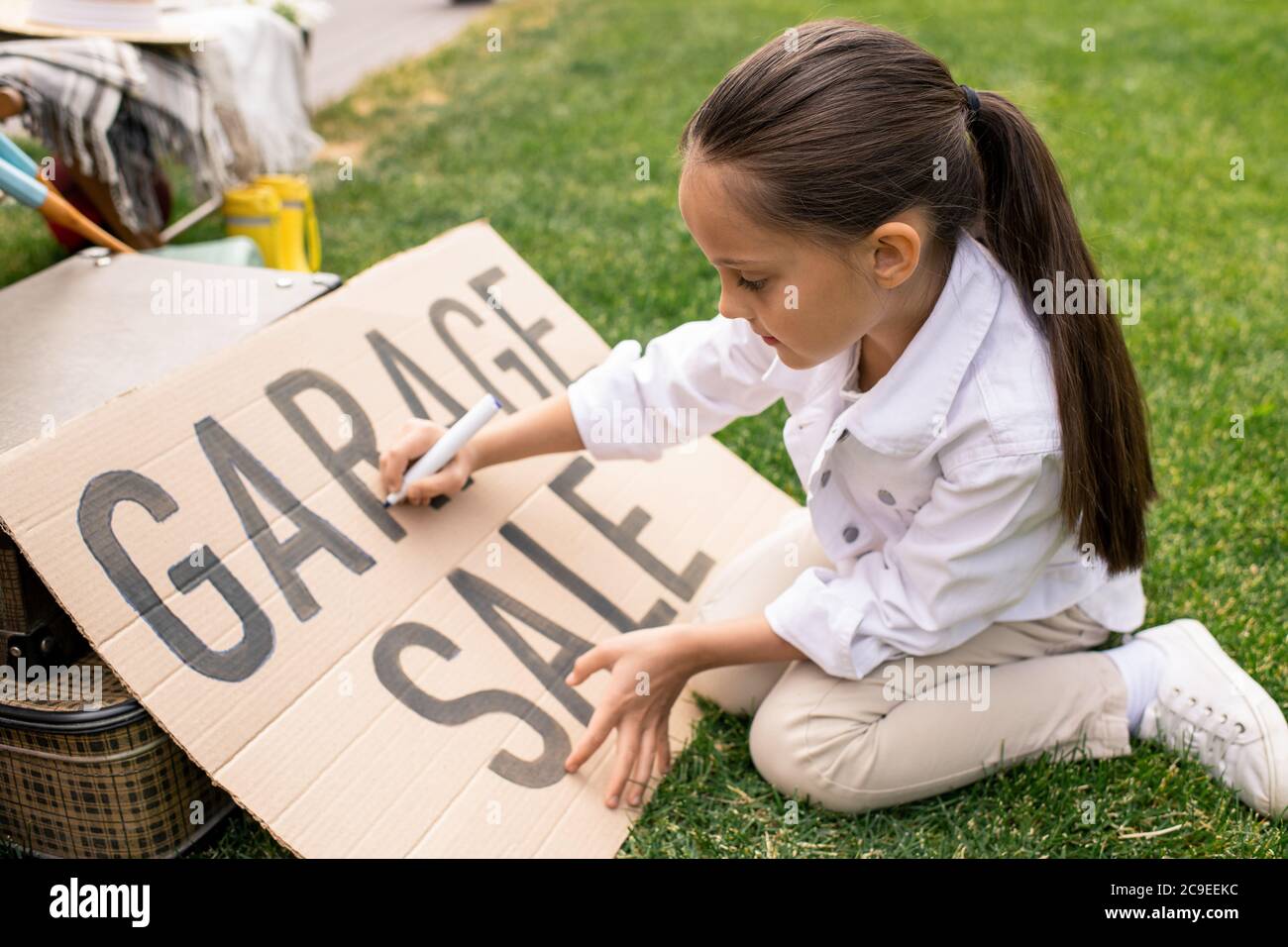 Hübsches Mädchen mit Pferdeschwanz sitzt auf Gras und macht Banner für Garage Verkauf im Hinterhof Stockfoto