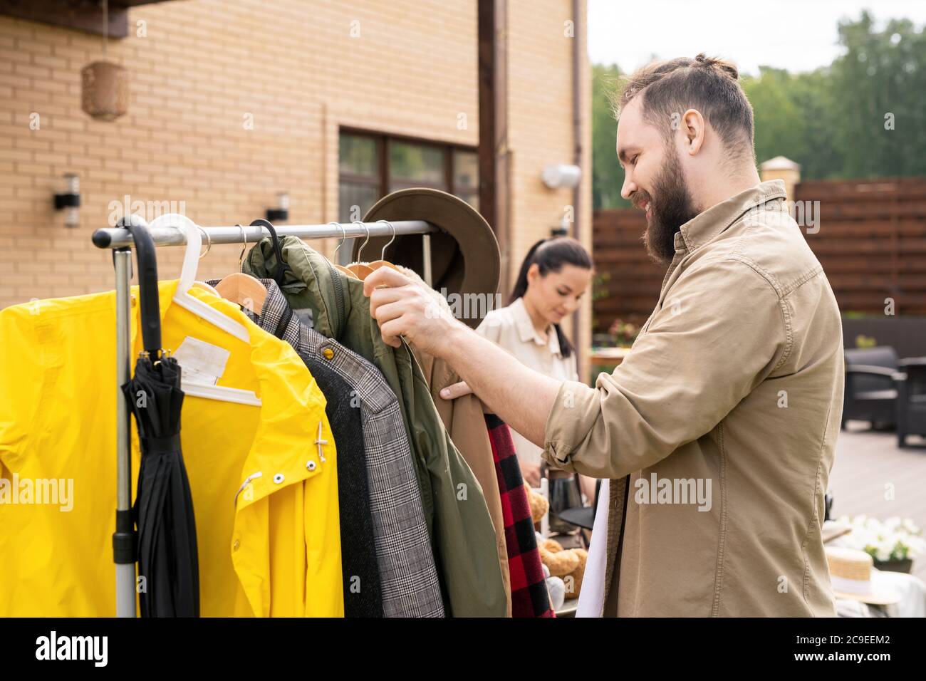 Lächelnder bärtiger Mann im Hemd, der am Regal steht und Kleidung beim Hofverkauf auswählt Stockfoto