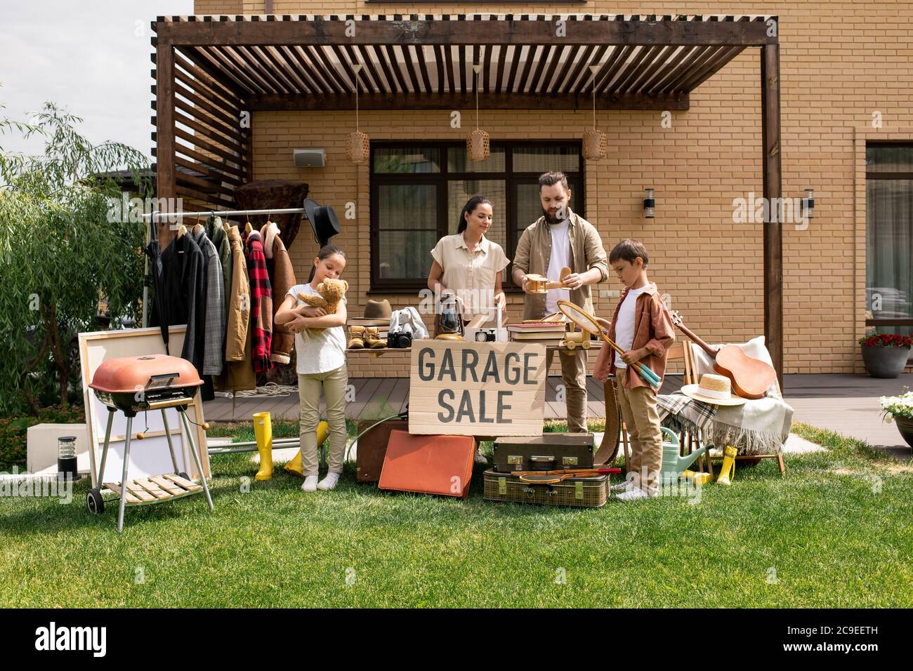 Junge Familie steht gegen Backsteinhaus und sagt Auf Wiedersehen zu alten Dingen im Garagenverkauf Stockfoto