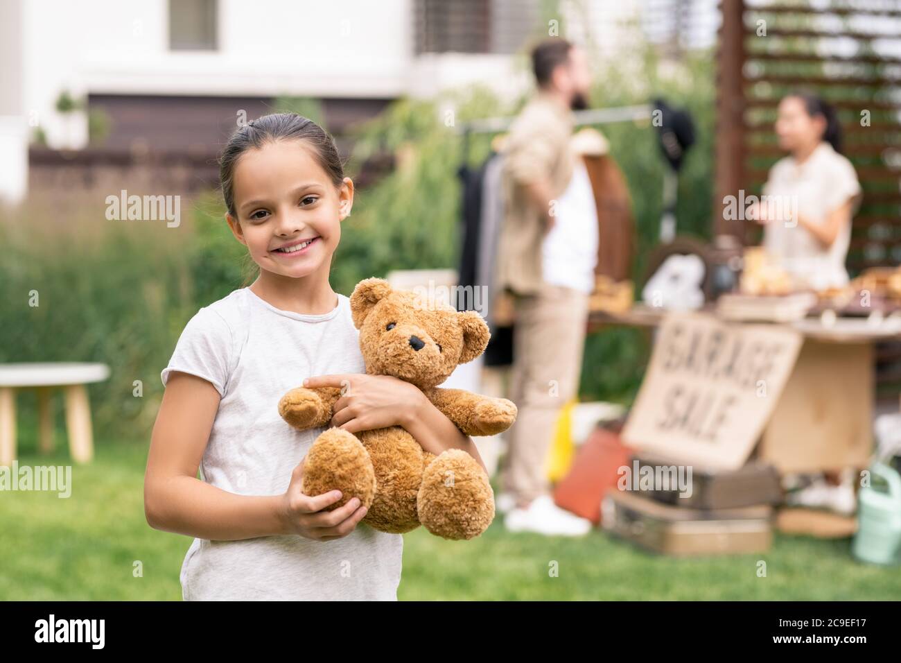 Portrait von fröhlichen hübschen Mädchen in weißen T-Shirt mit niedlichen Spielzeug Bär, sie besucht Garage Verkauf mit Vater Stockfoto