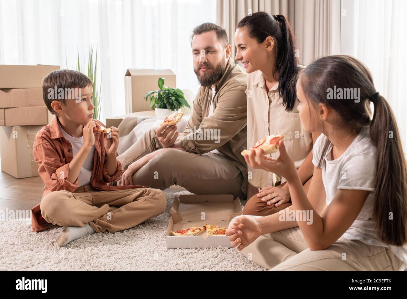 Freundliche junge Familie sitzt auf dem Teppich in einem neuen Haus voller Umzugskartons und Essen Pizza zum Abendessen Stockfoto