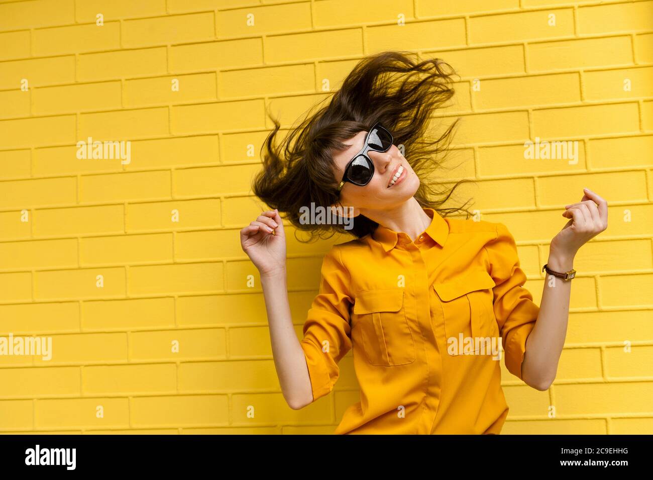 Ein Mädchen in einem gelben Hemd und in einer sonnengenähten Brille schüttelt den Kopf und ihr Haar entwickelt sich im Wind. Glückliches junges Mädchen in Sonnenbrille gegen einen Brut Stockfoto