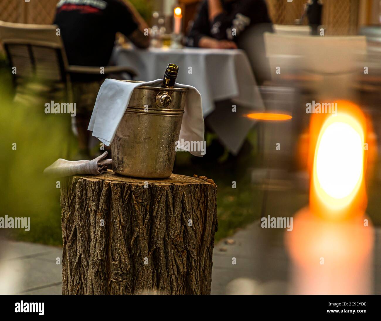 Ein Eiskübel mit einer gekühlten Flasche steht auf einem Baumstumpf im Außenbereich des Hotel Mühle in Binzen, Markgräflerland, Baden-Württemberg, Deutschland, während Gäste im Hintergrund bei Kerzenschein an einem gedeckten Tisch sitzen. Stockfoto