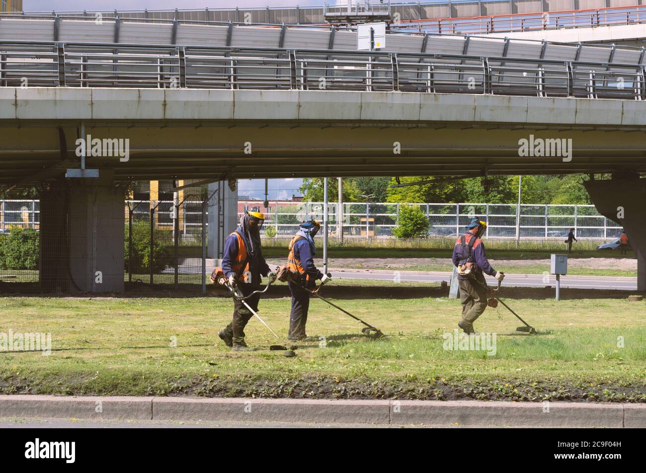 Sankt Petersburg, Russland - 26. Juli 2020: Drei Arbeiter mit Saitenbeschneidern in orangefarbenen Overalls mähen das Gras nahe der Autobahn Stockfoto