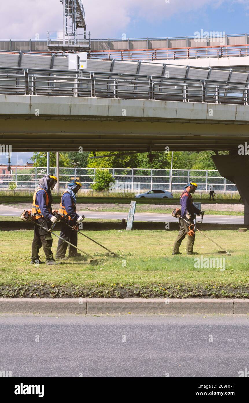 Sankt Petersburg, Russland - 26. Juli 2020: Drei Arbeiter in Overalls mähen das Gras auf dem Rasen mit Trimmer vertikale Ausrichtung Stockfoto