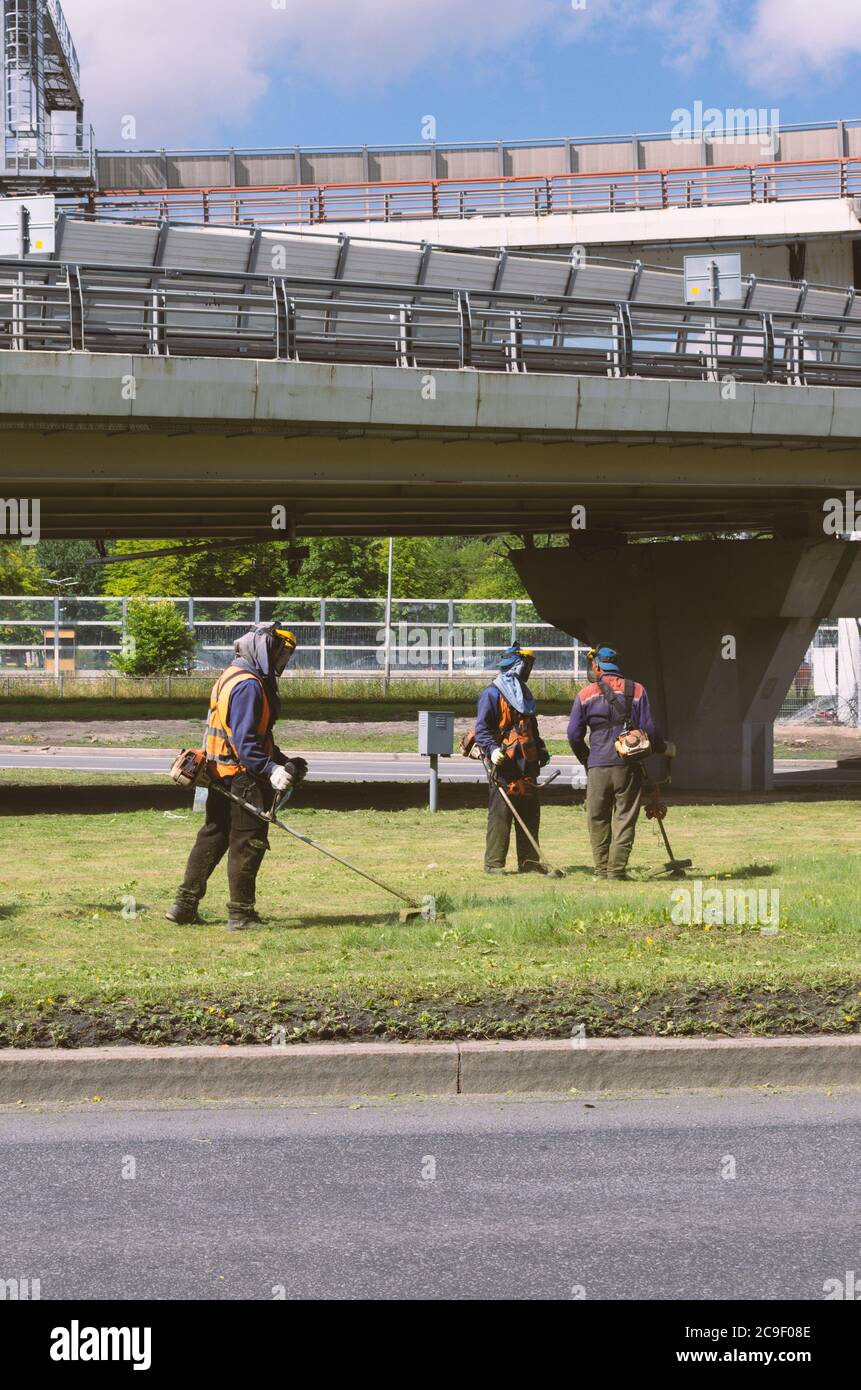 Sankt Petersburg, Russland - 26. Juli 2020: Kommunale Arbeiter in Aufruhr mähen das Gras mit Schnur Trimmer vor dem Hintergrund der Ringautobahn V Stockfoto