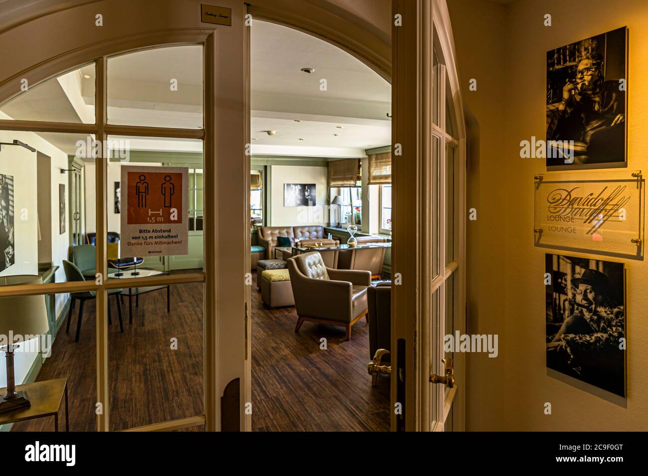 Ein Blick durch einen gewölbten Türbogen öffnet sich zur ehemaligen Raucherlounge im Hotel Mühle in Binzen, Markgräflerland, Baden-Württemberg, Deutschland – die Zigarrenlounge wurde neu gewidmet, und das Rauchen findet jetzt nur noch in den schwarz-weißen „Davidoff Lounge“-Fotografien an der Wand statt. Ein Schild bittet die Gäste, wegen der Coronavirus-Pandemie einen Abstand von 1,5 Metern einzuhalten. Stockfoto