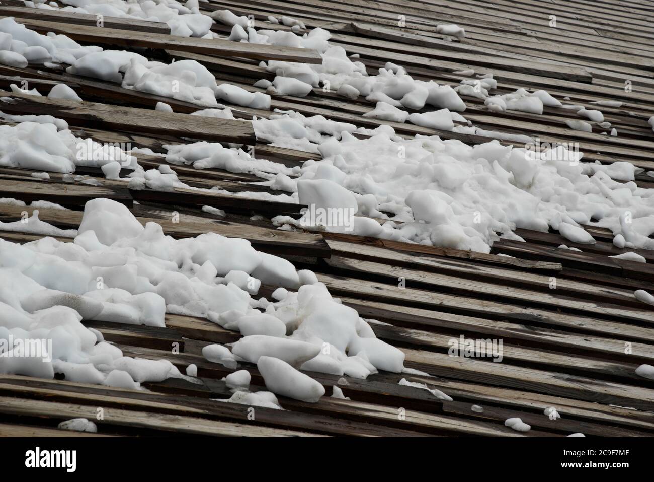 Auf einem alten Holzdach liegen Eis- und Schneeklumpen. Strahlend weißer Schnee im Kontrast zu den dunklen Holzlängen. Stockfoto