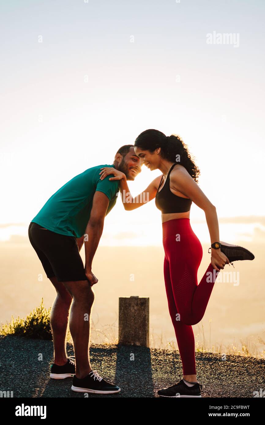 Fröhliches Paar, das morgens auf der Straße steht und sich aufwärmt. Porträt einer athletischen Frau, die ihre Beinmuskeln dehnt. Stockfoto