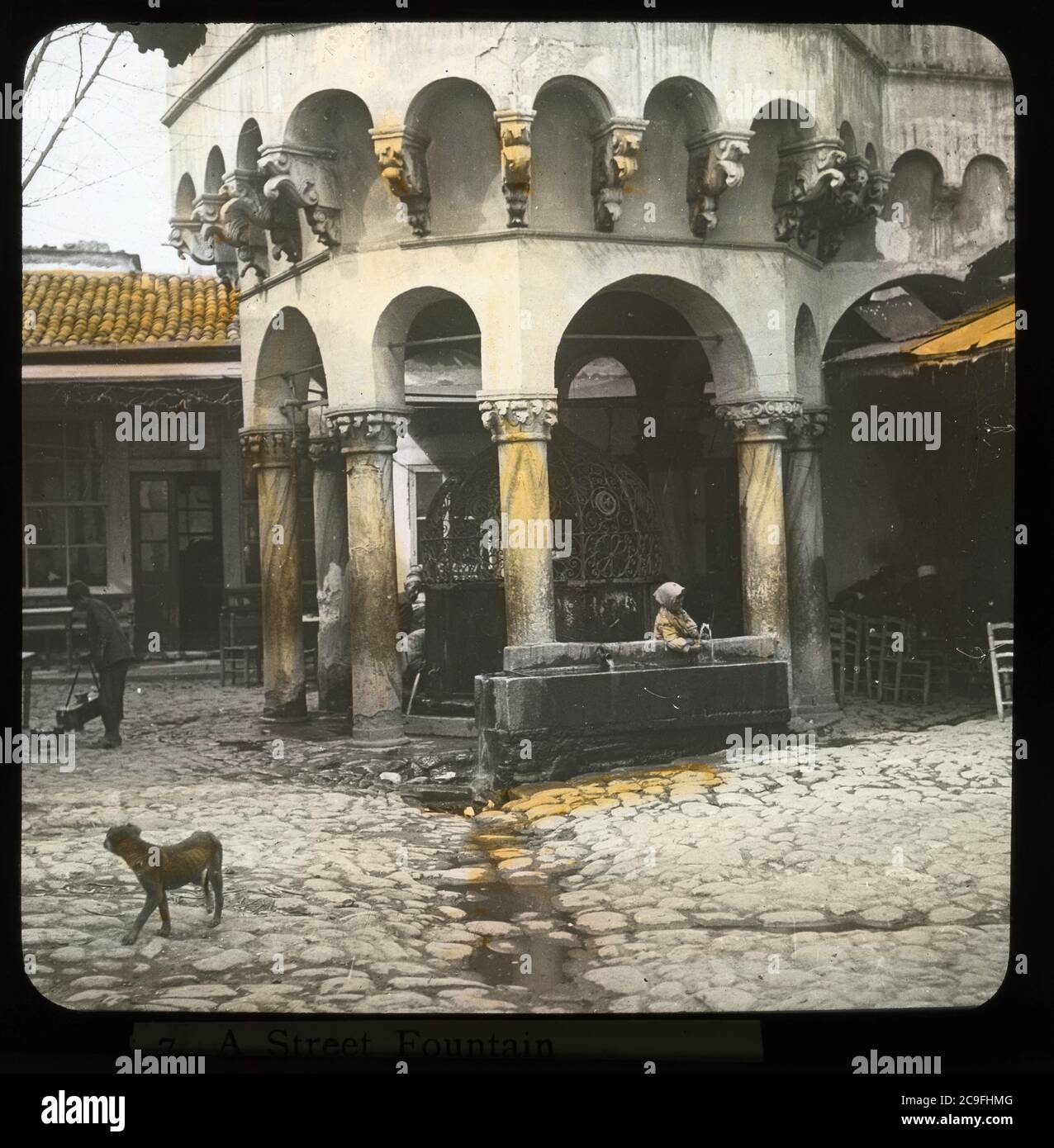 Şadırvan Camii Konak in Smyrna / Izmir, Türkei. Frau, die Wasser aus einem Wasserplatz neben dem Şadırvan zieht. Magerer Hund und Händler in der Nähe. Handkoloriertes Foto auf trockenem Glasplatte aus der Sammlung Herry W. Schaefer, um 1910. Stockfoto