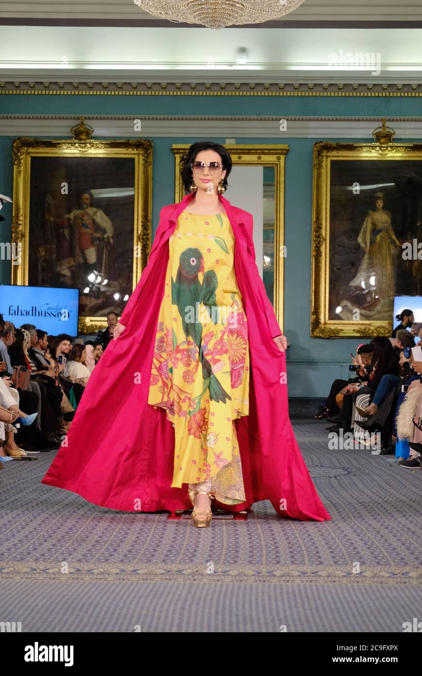 Model trägt einen langen roten Mantel und gelbes Outfit mit einem Papagei Design Spaziergänge für den pakistanischen Designer Fahad Husseyn in London. Stockfoto