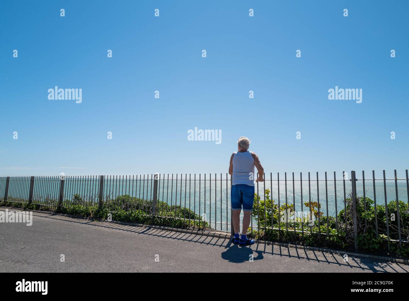 Ein älterer Mann, der ein Trägeroberteil und Jeans-Shorts trägt, blickt an einem Sommertag über Geländer auf einer Promenade zum Meer. Stockfoto