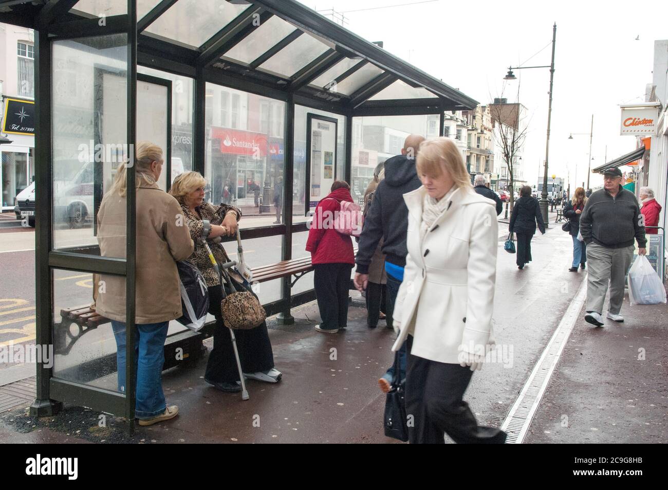 Auf Bexhill in der Sea High Street gehen viele Leute einkaufen, andere Leute stehen an der Bushaltestelle. Stockfoto