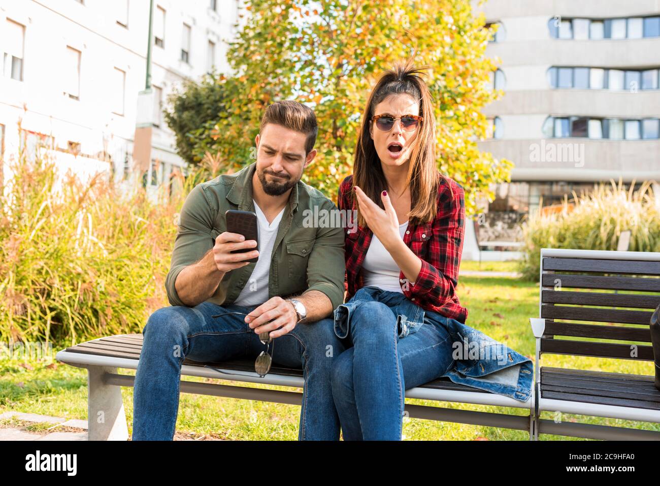 Ein Paar ist schockiert über Inhalte auf dem Bildschirm eines Telefons gesehen, während im Sommer auf einer Parkbank in der Stadt sitzen. Stockfoto