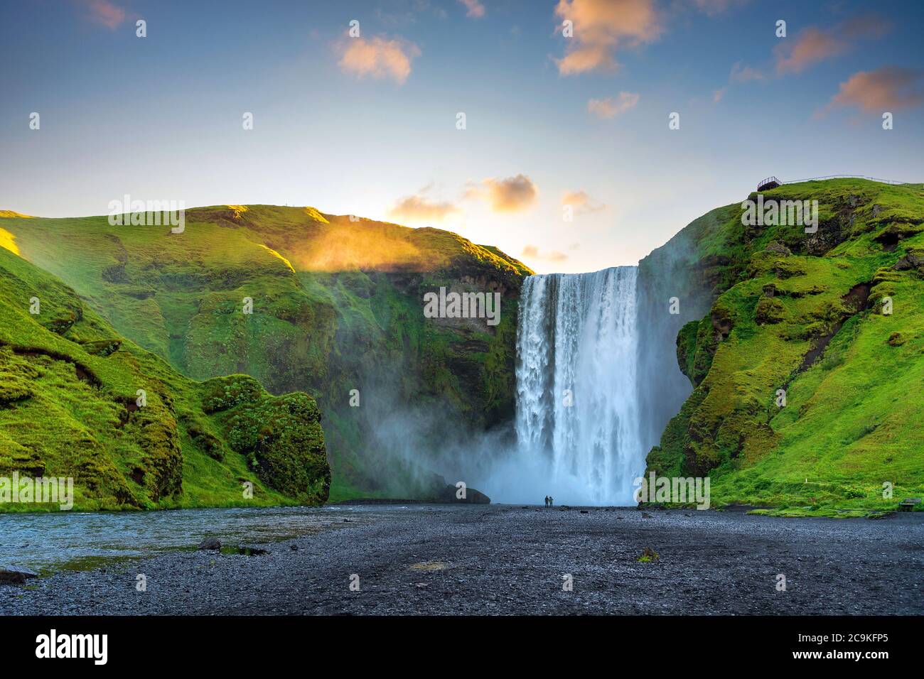 Männer und Frauen stehen und beobachten die großen Skogafoss Wasserfälle. Am Morgen kommt der Sonnenaufgang hinter den Bergen, mit grünem Gras all o Stockfoto