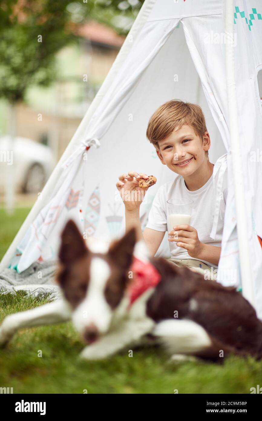 Der kleine Junge genießt das Essen mit seinem Hund in einem Camp an einem sonnigen Tag Stockfoto