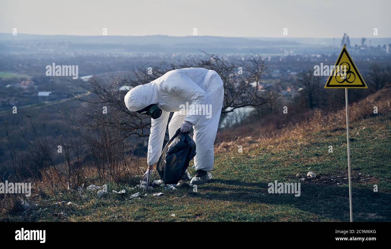 Ökologe trägt weißen Schutzanzug, Gasmaske, sammeln Plastikmüll in schwarzen Abfallbeutel im Freien an einem sonnigen Tag, Biohazard Symbol auf der rechten Seite. Konzept der Umweltverschmutzung Stockfoto