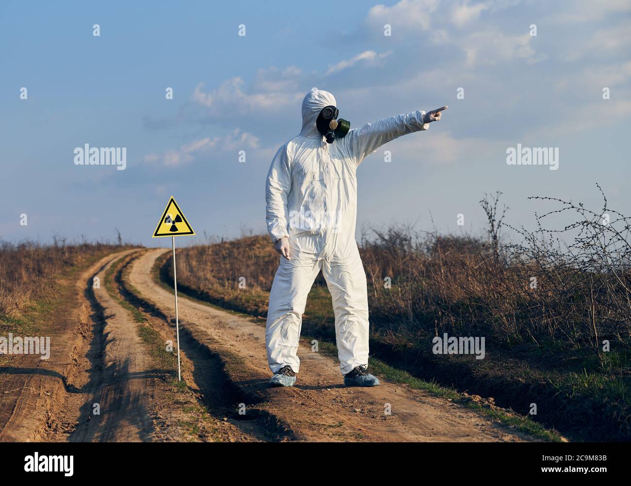 Mann in weißem Overall, Gasmaske und Handschuhe stehen auf Dorfstraße auf dem Feld neben Strahlungszeichen zeigen und wegschauen, an sonnigen Tag unter blauem Himmel. Konzept der Ökologie, biologische Gefahr. Stockfoto