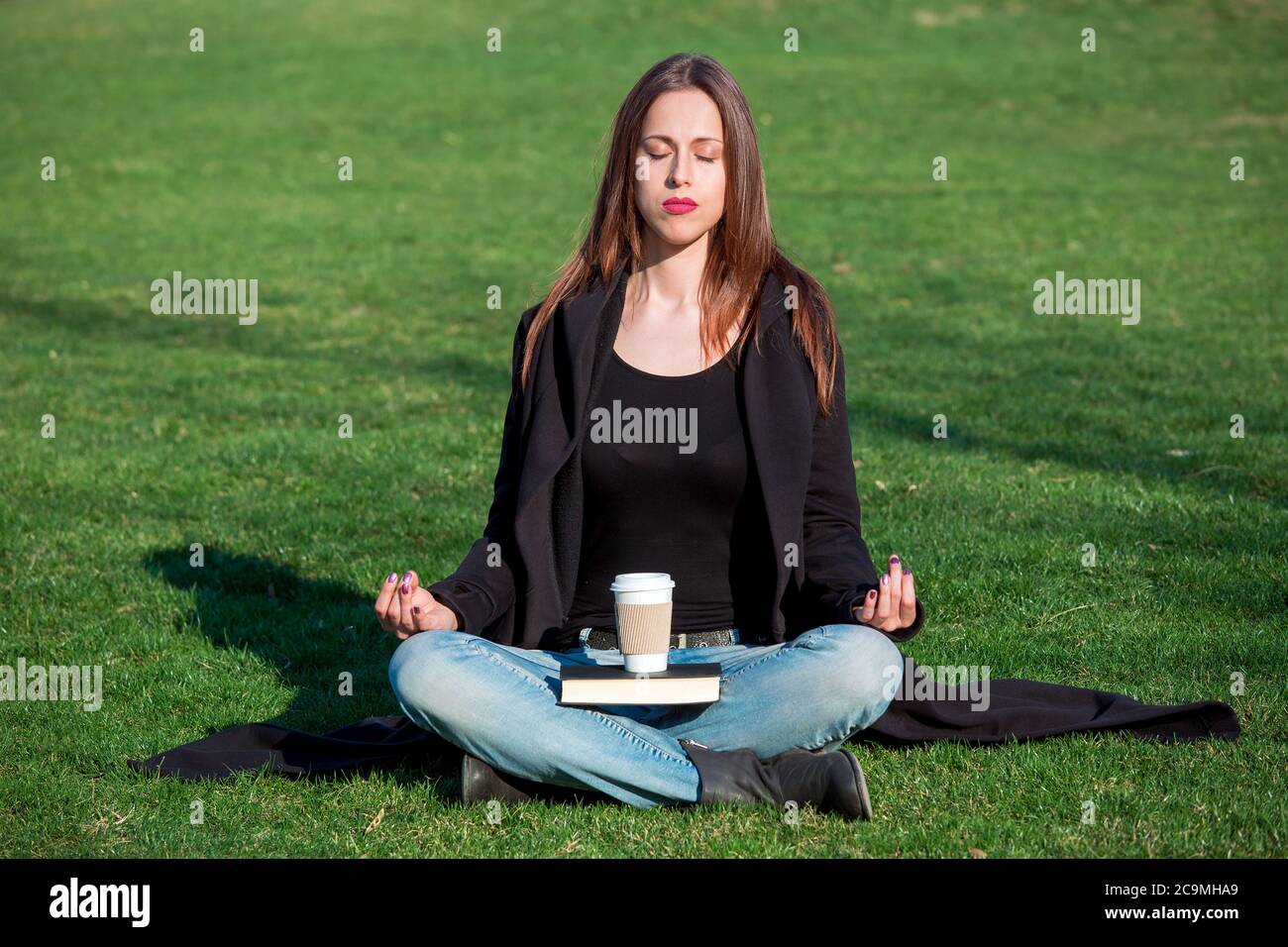 Das Mädchen sitzt mit geschlossenen Augen in der Lotusposition und  meditiert an einem sonnigen Tag auf dem Gras im Park, in einem schwarzen  Mantel und blauen Jeans auf ihr Stockfotografie - Alamy