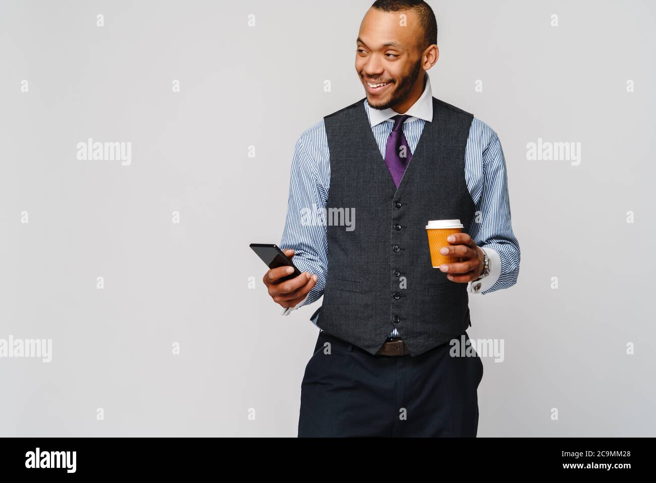 Junger afroamerikanischer Geschäftsmann hält Telefon und Kaffee über hellgrauen Hintergrund zu gehen Stockfoto