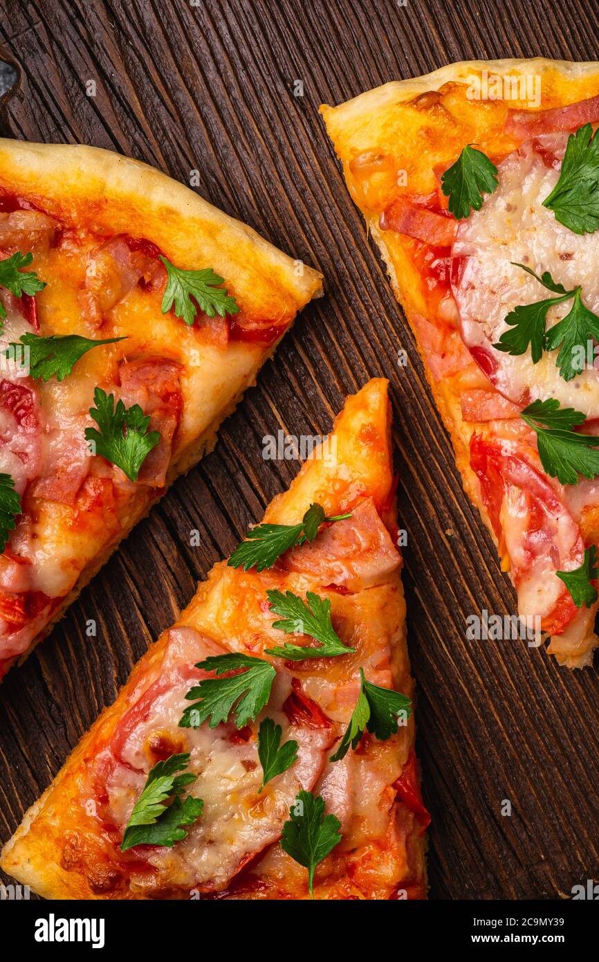 Heiße Pizzascheiben mit Mozzarella-Käse, Schinken, Tomate und Petersilie auf braunem Holzschneidebrett, Draufsicht Makro Stockfoto