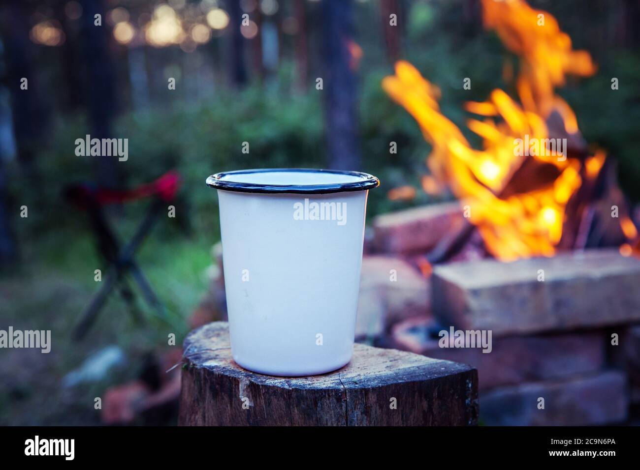 Nahaufnahme von Metall Camping Cup, auf Holz Stumpf, mit Lagerfeuer und Camping Stuhl im Hintergrund. Stockfoto