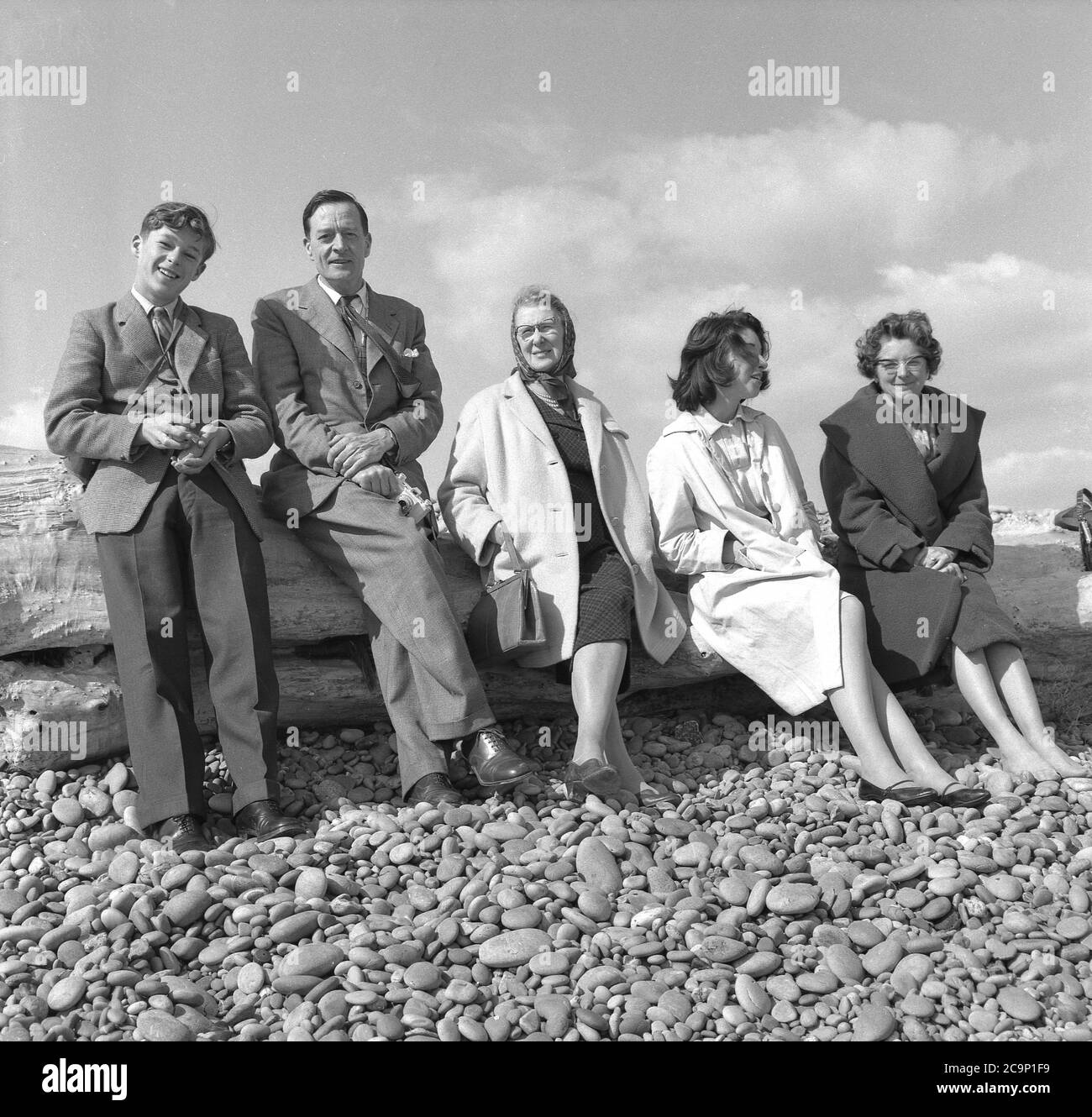 1950s, historisch, eine britische Familie, die auf einem Baumstamm an einem Kiesstrand ruht, alle formal gekleidet, der Mann in Anzügen, der junge Mann eine Jacke und Krawatte und die Frauen alle in langen Mänteln, die in dieser Zeit war normale Alltagskleidung, auch wenn Sie die Küste besuchen, England, Großbritannien. Stockfoto