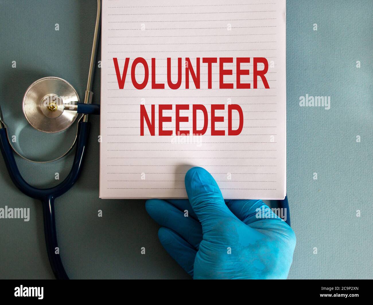 Hand in blauem Handschuh, weiße Karte mit den Worten "Freiwilliger benötigt" und Stethoskop. Medizinisches Konzept. Stockfoto