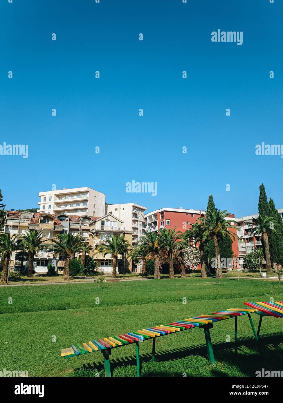 Jugoslawische Häuser im Zentrum von Budva in Montenegro, neben dem Stadtpark und grünen Dattelpalmen, mit einer grünen Wiese und blauem Himmel im Sommer. Stockfoto