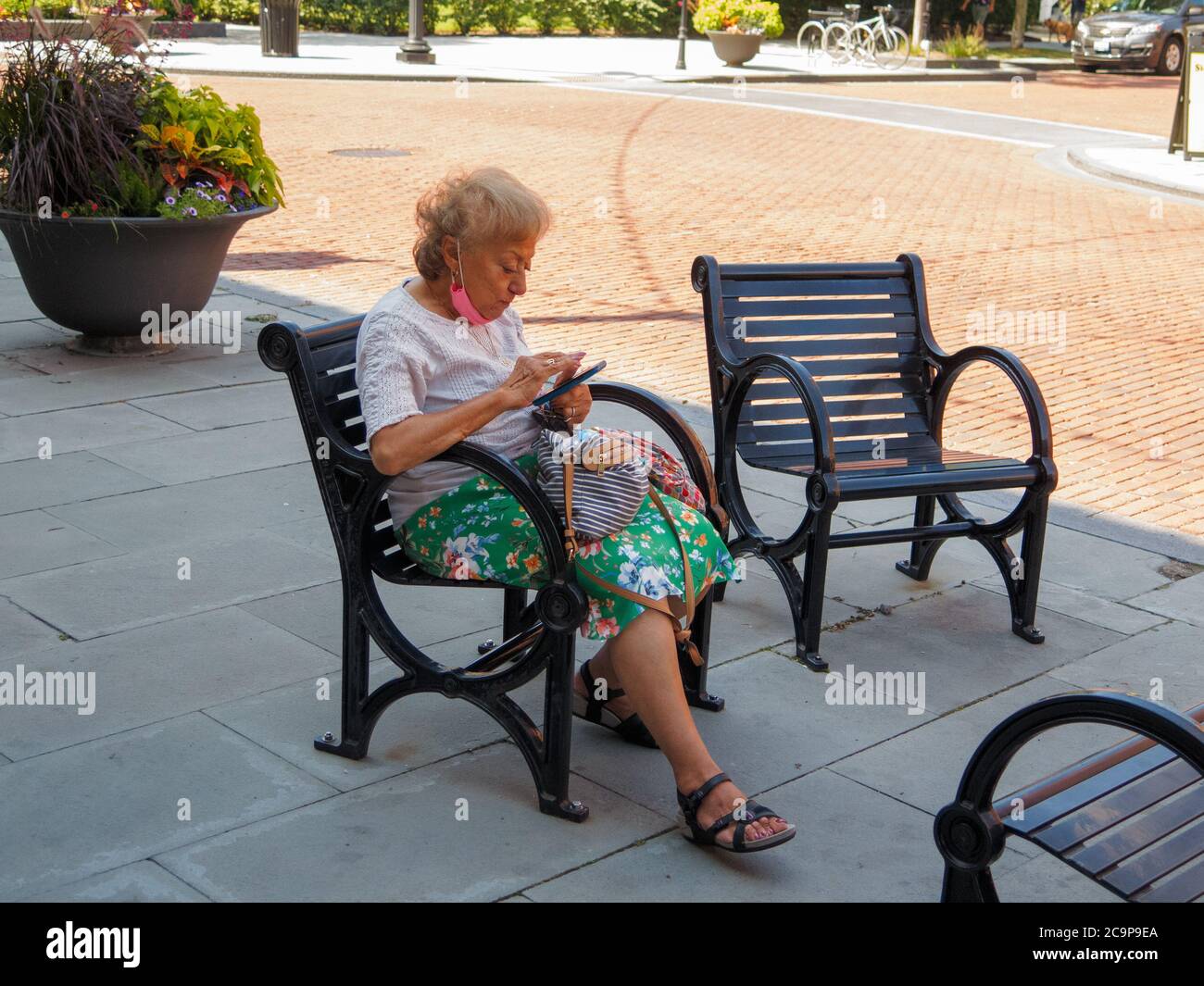 Frau, die auf einem öffentlichen Stuhl mit dem Mobiltelefon sitzt. Gesichtsmaske unter dem Kinn gezogen. Stockfoto