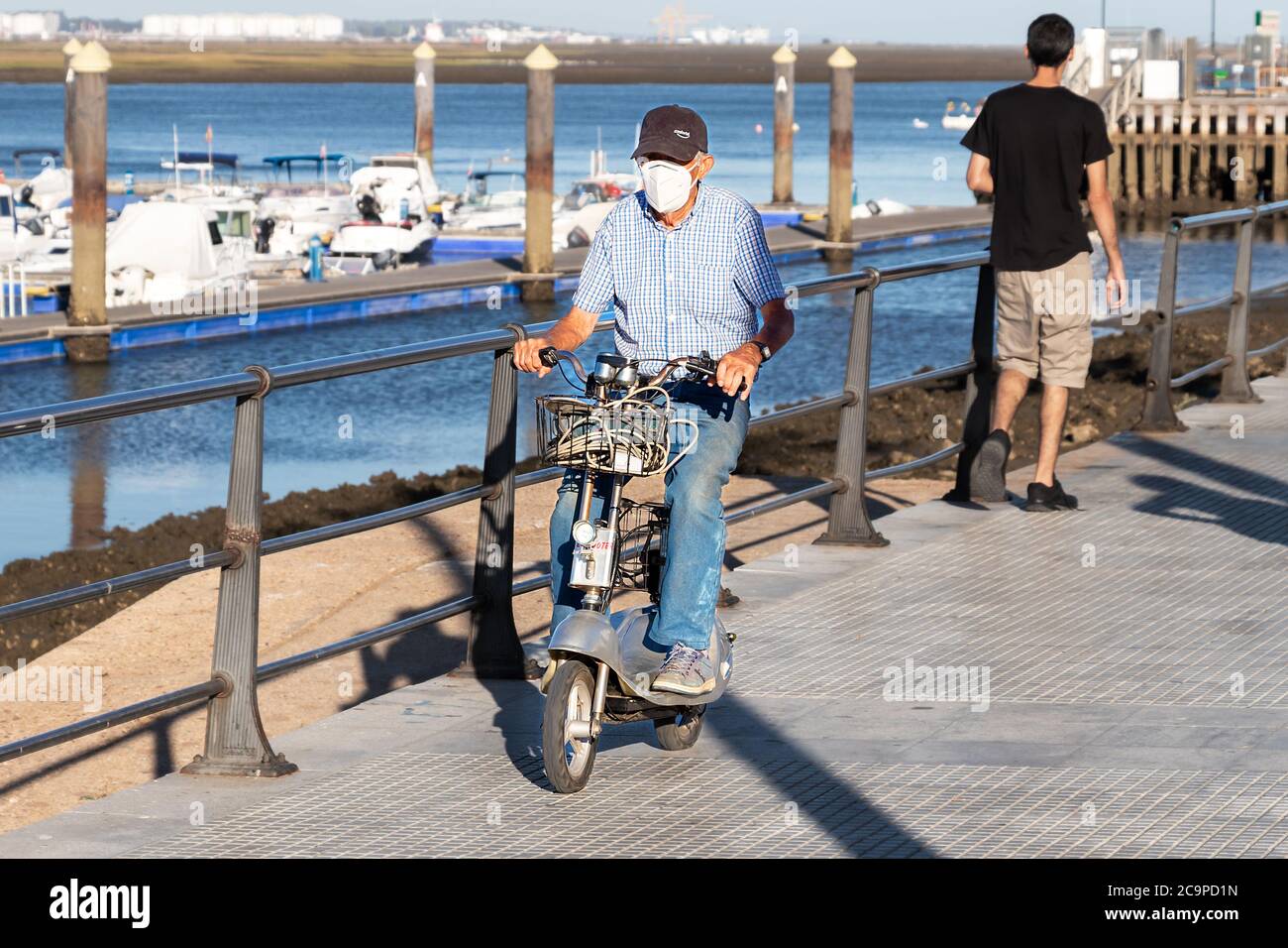 Punta Umbria, Huelva, Spanien - 10. Juli 2020: Ein älterer Mann, der mit einer Schutzmaske auf dem Bürgersteig einen Elektroroller fährt. Stockfoto