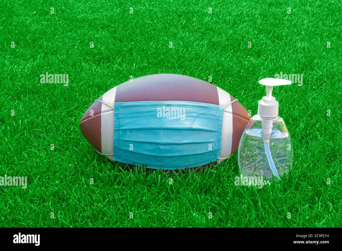 Ein American Football mit Gesichtsmaske und Händedesinfektionsmittel rechts auf dem Spielfeld mit grünem Gras. Konzept Fußball während Pandemie Stockfoto