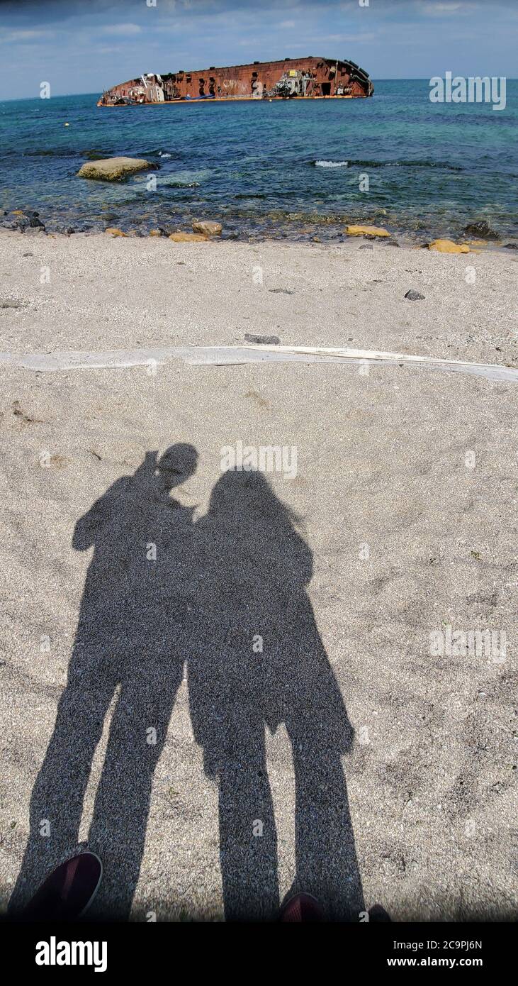 Schattenbilder von Mann und Frau auf Strandsand und altes rostig zerstörtes Schiff im Wasser auf verschwommenem Hintergrund Stockfoto
