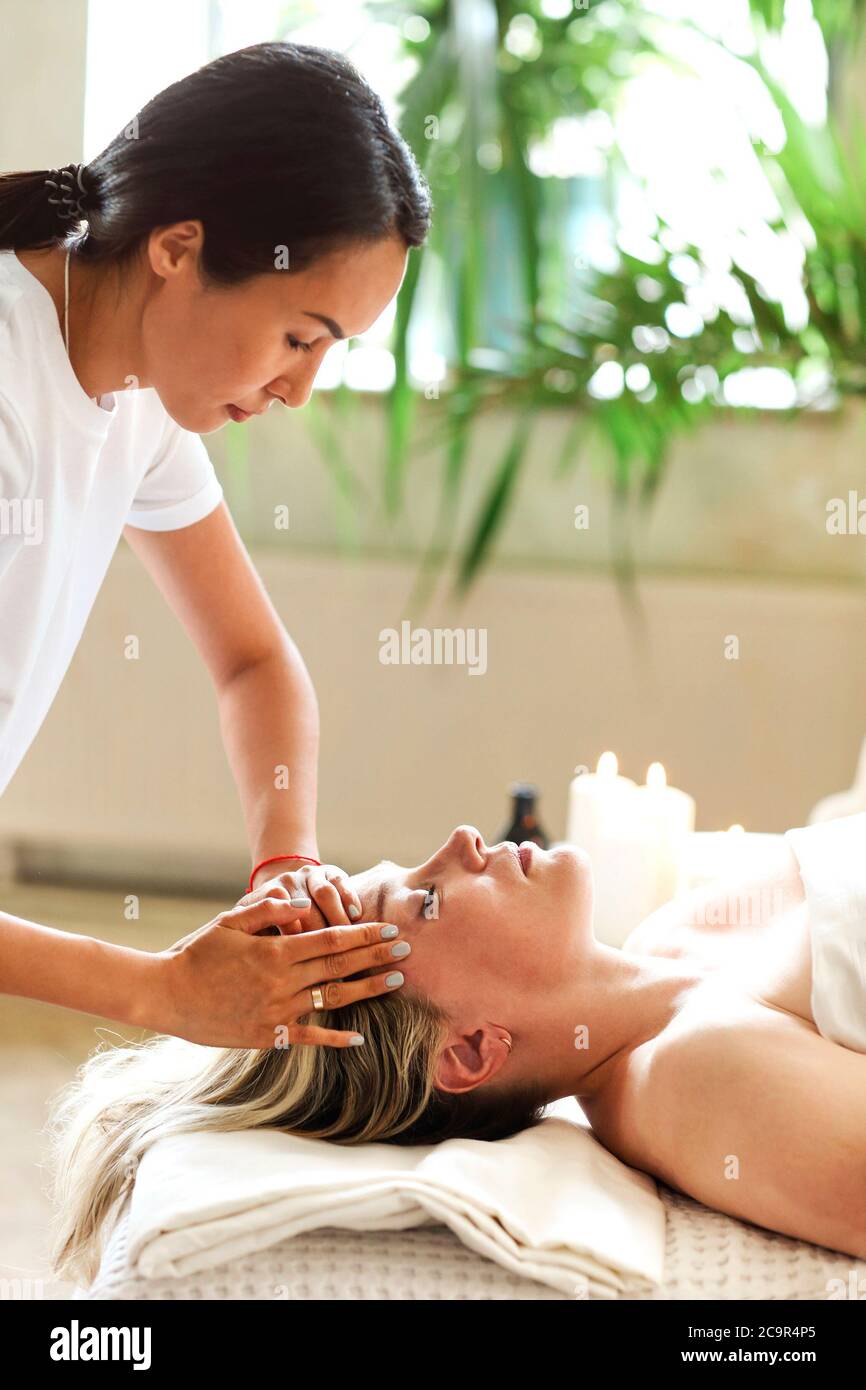 Seitenansicht der ethnischen weiblichen Therapeutin, die das Gesicht einer Frau mittleren Alters massiert, die auf dem Massageliegen im Spa-Salon liegt Stockfoto