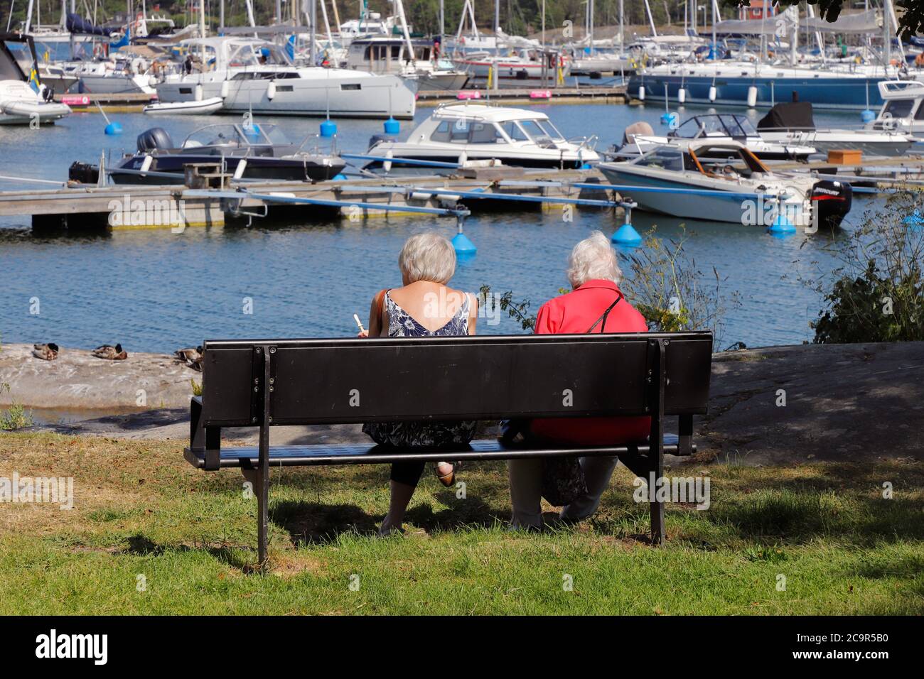 Nynashan, Schweden - 26. Juni 2020: Zwei ältere Frauen sitzen auf einer Parkbank mit der Marina im Hintergrund Stockfoto