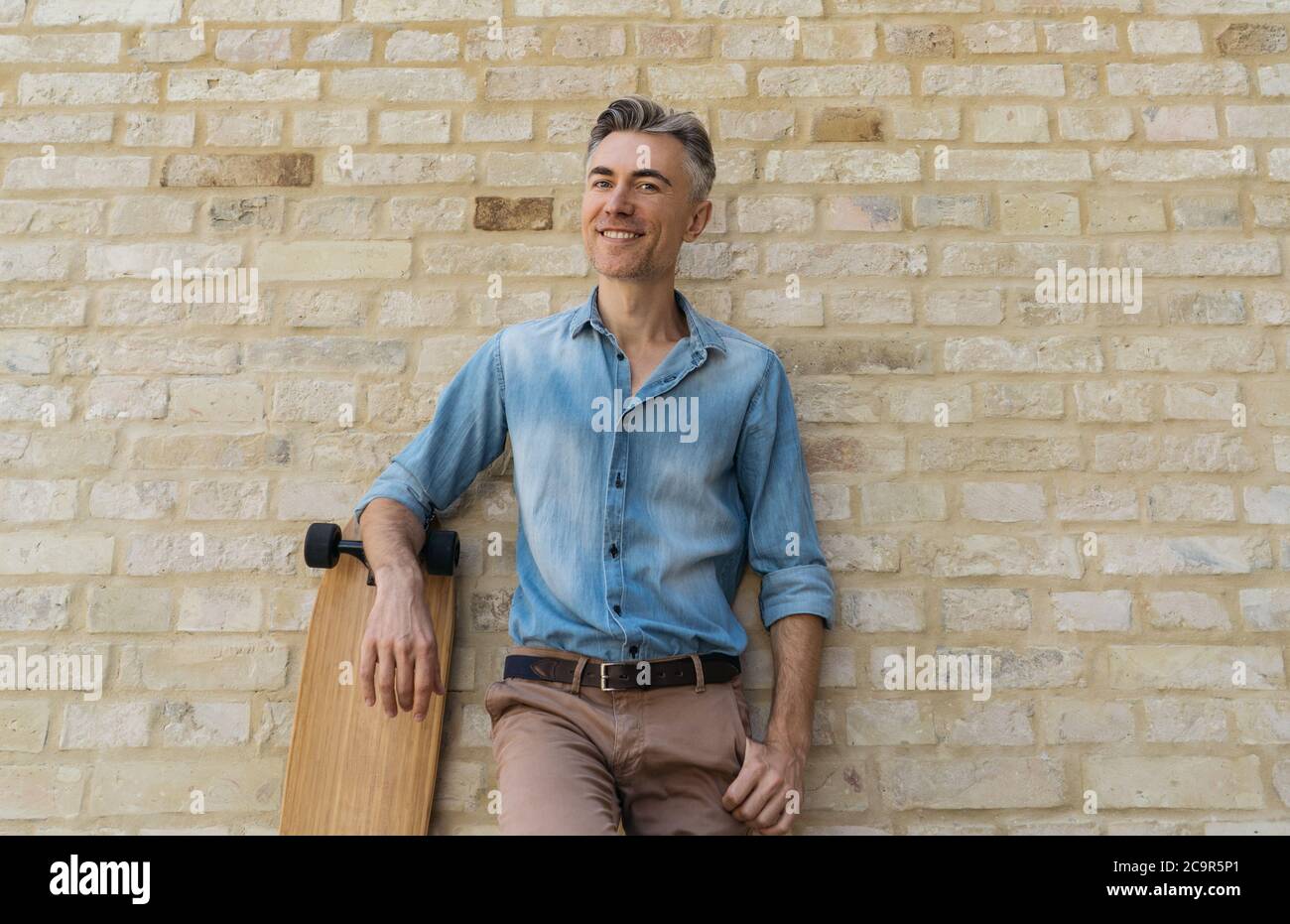 Stilvolle reife Mode-Modell tragen lässige Kleidung, posiert für Bilder. Fröhlicher Skater mit Longboard, steht an der Wand Stockfoto