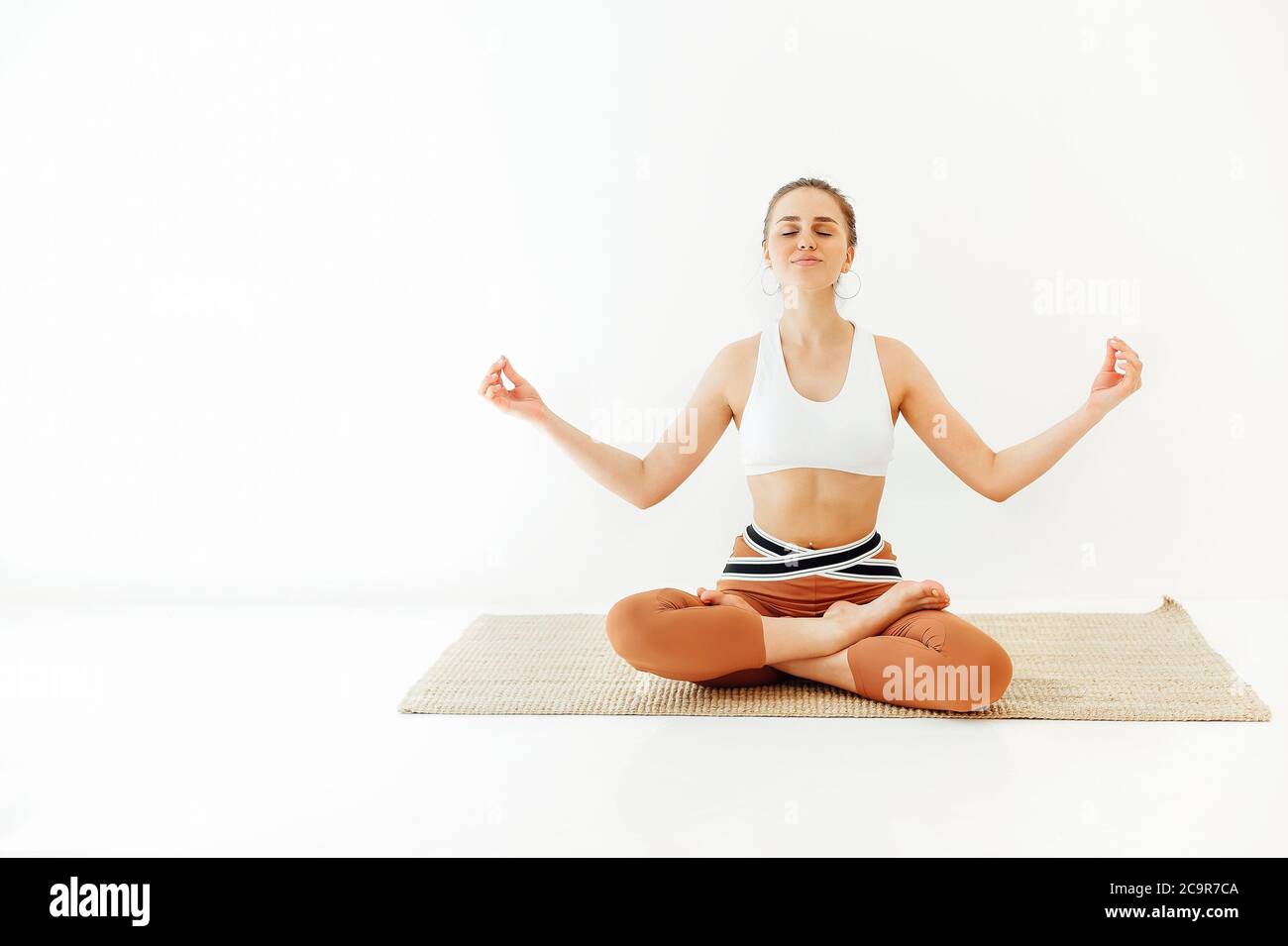 Junge Frau in Sportbekleidung sitzt mit gekreuzten Beinen und Mudra-Händen auf Matte und macht Yoga während Achtsamkeit üben Stockfoto