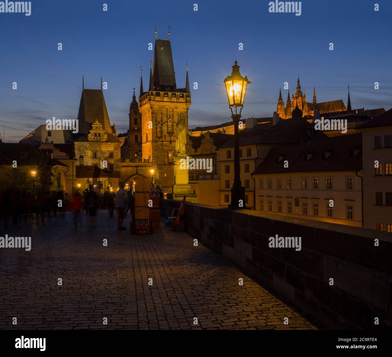 Blick von der Karlsbrücke auf den Mala Strana-Brückenturm mit glühender Straßenlampe nad Prager Burg bei Nacht, dunkelblauer Himmel. Stockfoto