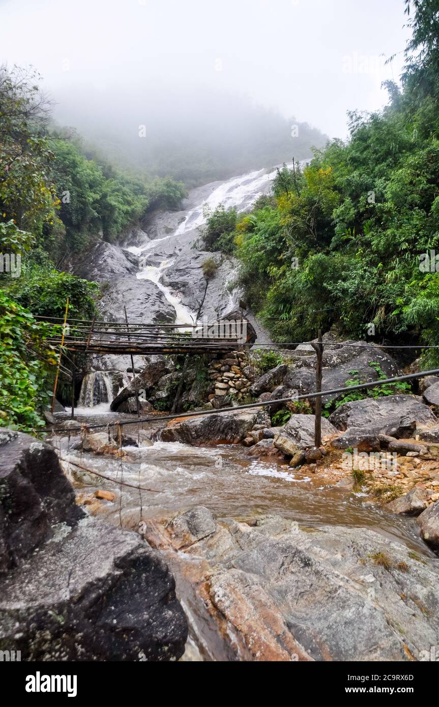 Ein Bach mit einem rauschenden Wasserfall fließt einen felsigen und felsigen Hang hinunter. Eine kleine Holzbrücke überspannt den Fluss. Neblige Situation in Sapa, Vietnam. Stockfoto