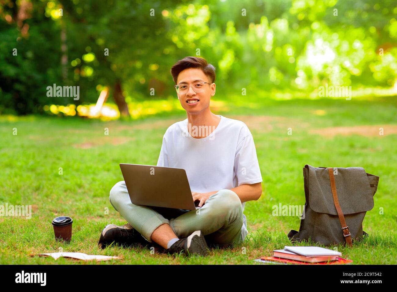 Lächelnder asiatischer Student Kerl sitzt mit Laptop im Freien, Vorbereitung für Prüfungen Stockfoto