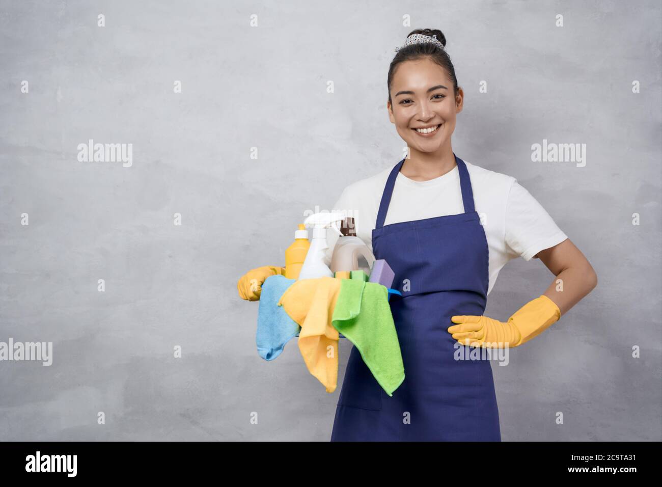 Porträt einer glücklichen jungen Frau, Putzfrau in Uniform mit Eimer von Reinigungsmitteln, die an der grauen Wand stehen und die Kamera anlächeln. Studioaufnahme. Housekeeping und Reinigungsservice Konzept Stockfoto