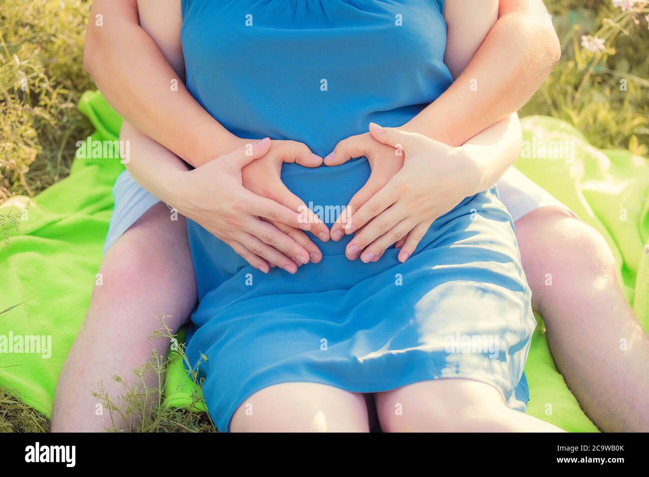 Schwangere Frau und ihr Mann sitzen im Park und halten ihre Hände in Herzform auf ihrem Baby Beule. Nahaufnahme. Familie, Schwangerschaft, Liebe concep Stockfoto