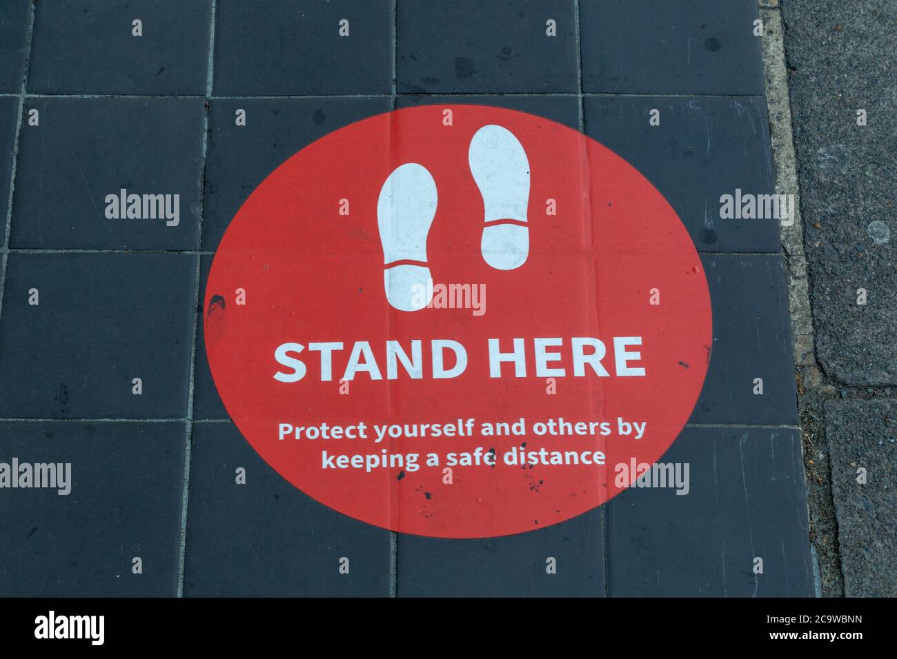Ein Schild auf dem Boden vor einem Geschäft, auf dem Kunden mitgeteilt wird, wo sie Schlange stehen sollen, damit sie sich gemäß den Richtlinien der Regierung sozial distanzieren Stockfoto