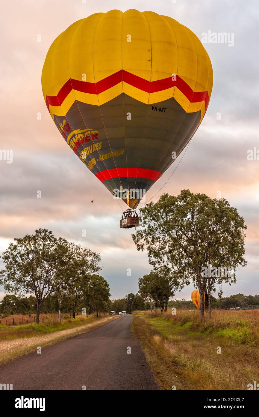 Ein farbenfroher Ballon mit Touristen beginnt sein Abenteuer im Morgengrauen, das langsam über Bäumen und einer Landstraße in Mareeba, Queensland, Australien, aufsteigt. Stockfoto